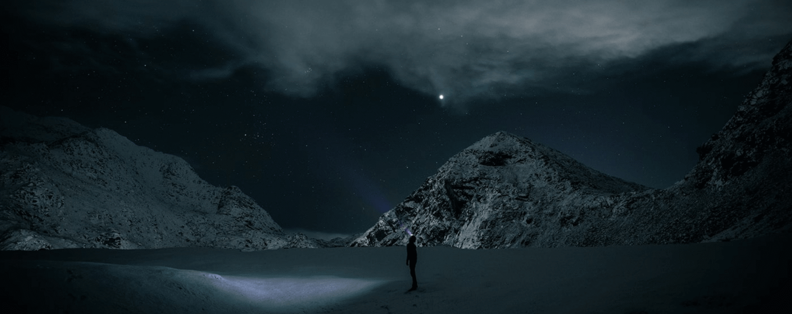 Uttakleiv Strand bei Nacht im Winter: eine Person steht allein im Schnee am Wasser, dahinter schneebedeckte Berge unter dem Sternenhimmel der Lofoten