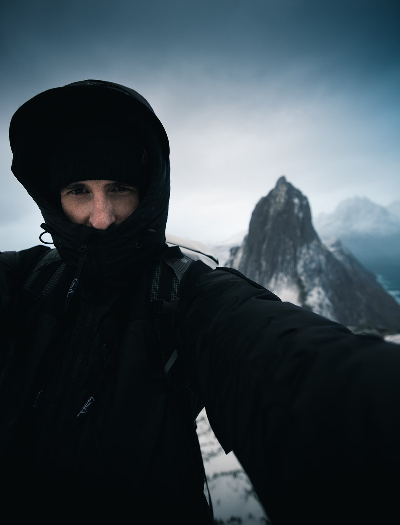Selfie mit warmer Winterjacke, Mütze und eng zugezogener Kapuze bei arktischem Sturm auf Senja, im Hintergrund der Gipfel des Segla