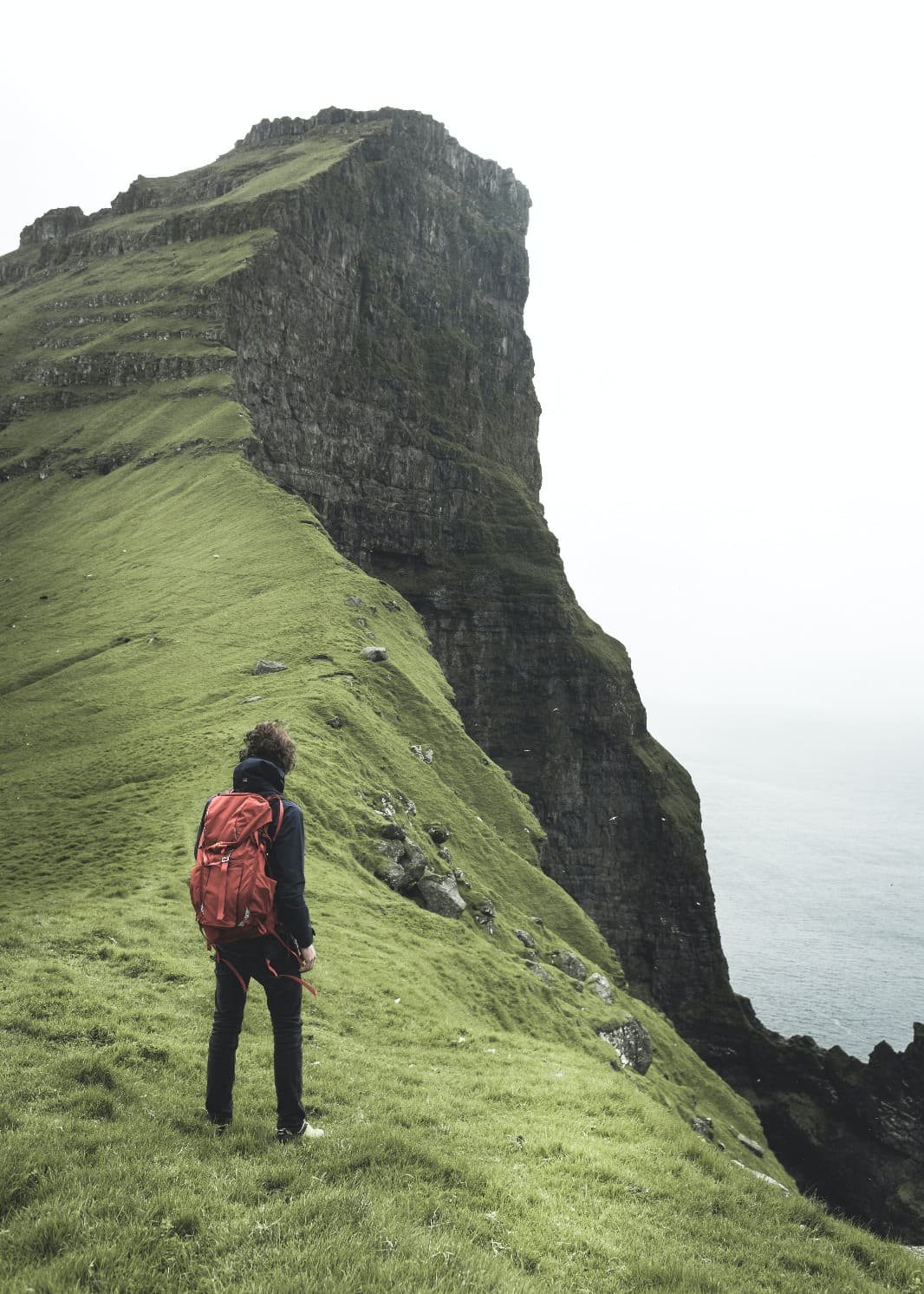 Wanderer mit rotem Rucksack vor den steilen Klippen am Nordende von Kalsoy