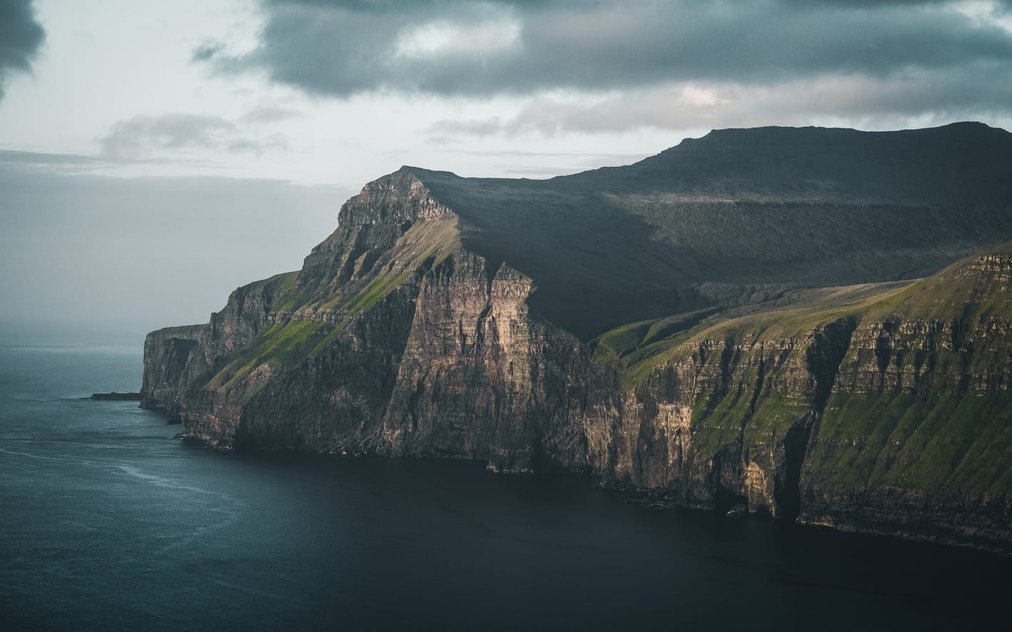 Steile, grün bewachsene Klippen der Färöer-Inseln über dem Nordatlantik unter bewölktem Himmel.