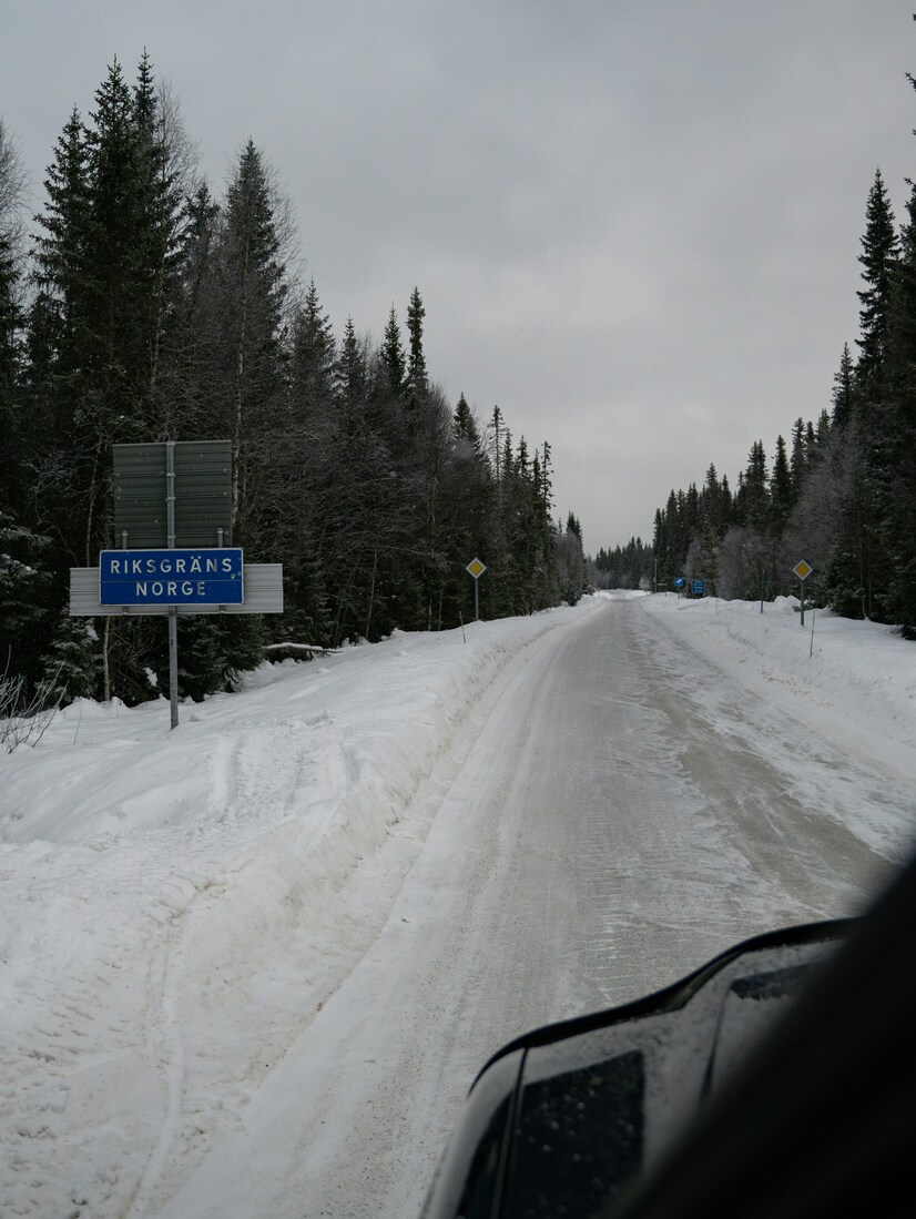 Straße mit Straßenschild ›Riksgräns Norge‹, aus der Perspektive eines Autofahrers fotografiert.