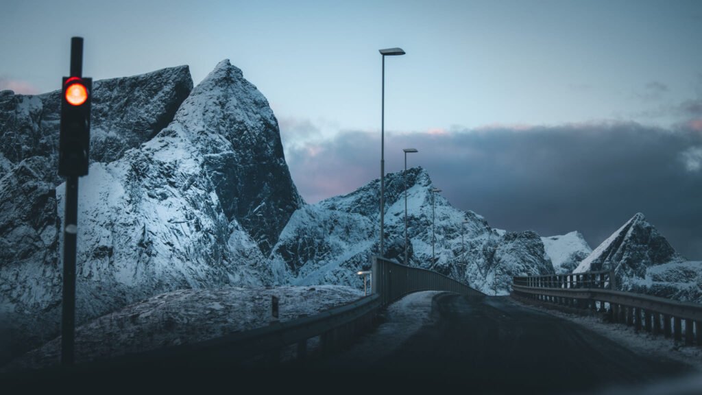 Verschneite Berge und eine Brücke mit Straßenlaternen in der blauen Stunde in Nordnorwegen, rote Ampel im Vordergrund