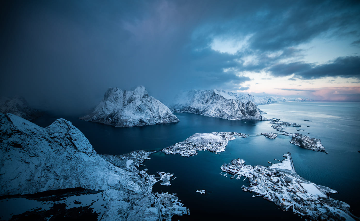 Blick vom Reinebringen über das verschneite Reine und Hamnøy, dunkler Fjord und Schneeschauer über den Bergen der Lofoten