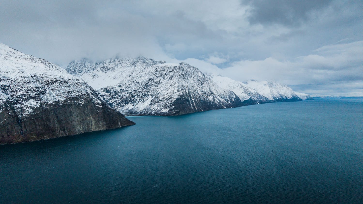 Drohnenaufnahme der Lyngen Alps, deren schneebedeckte Steilwände direkt aus dem Fjord aufsteigen, unter tiefer Wolkendecke