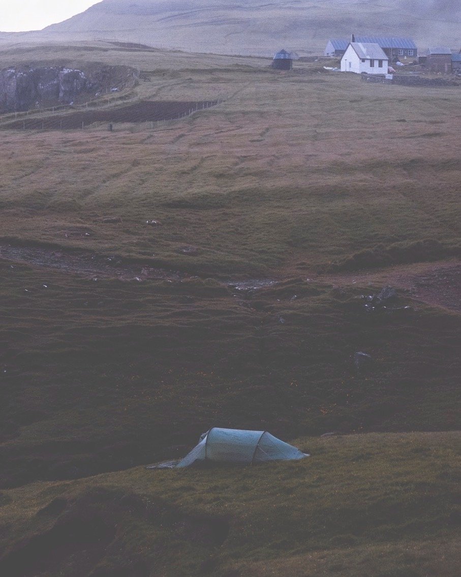 Kleines Zelt am Hang in Gásadalur in der Dämmerung, im Hintergrund die Häuser des Dorfes und die Berge im Dunst