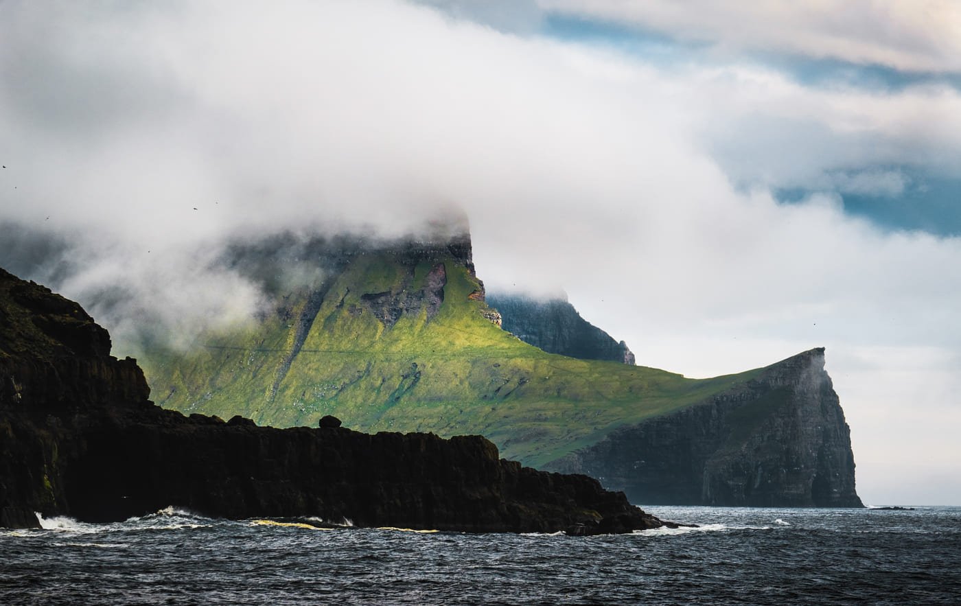 Steilküste von Vágar mit Wolken über der Bergkante, im Wasser die dunklen Basaltklippen und rechts hinten das Felsentor von Drangarnir