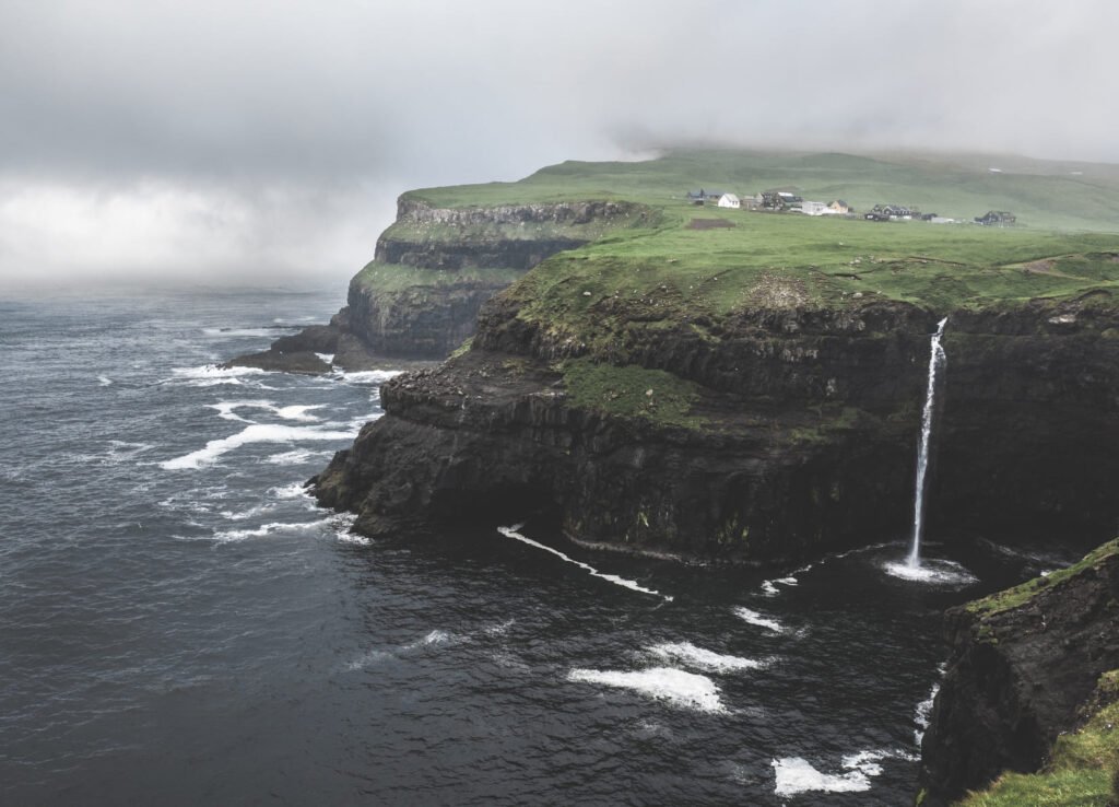 Der Wasserfall Múlafossur stürzt bei bedecktem Himmel von der Klippe ins Meer, dahinter das Dorf Gásadalur auf der Insel Vágar