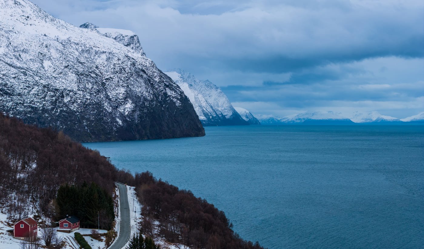 Blick vom Westufer des Lyngenfjords nach Norden, links die schneebedeckten Lyngen Alps, im Vordergrund eine rote Hütte und die Küstenstraße am Hang