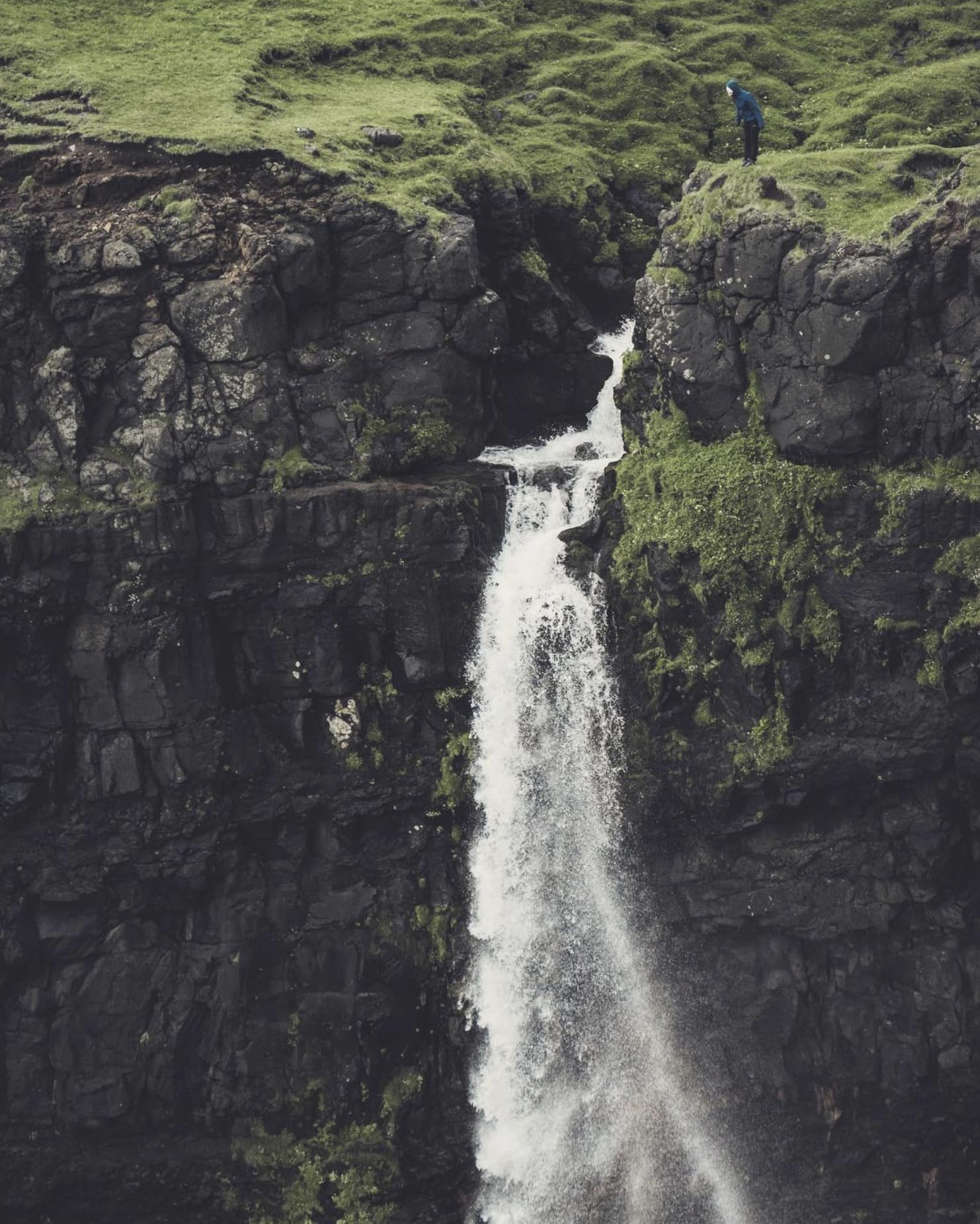Der Wasserfall Múlafossur stürzt über die dunkle Basaltkante, rechts oben steht eine Person am Rand der Klippe